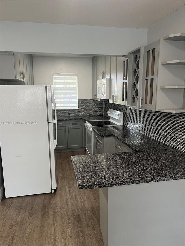 kitchen with white appliances, dark wood-type flooring, a peninsula, dark stone countertops, and decorative backsplash