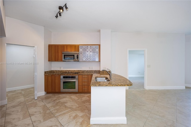 kitchen featuring a peninsula, dark stone countertops, a sink, stainless steel appliances, and brown cabinetry
