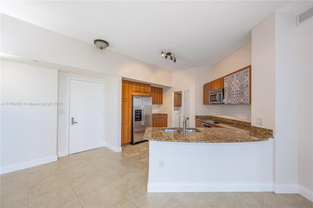kitchen featuring appliances with stainless steel finishes, brown cabinets, washer and dryer, dark stone counters, and a sink