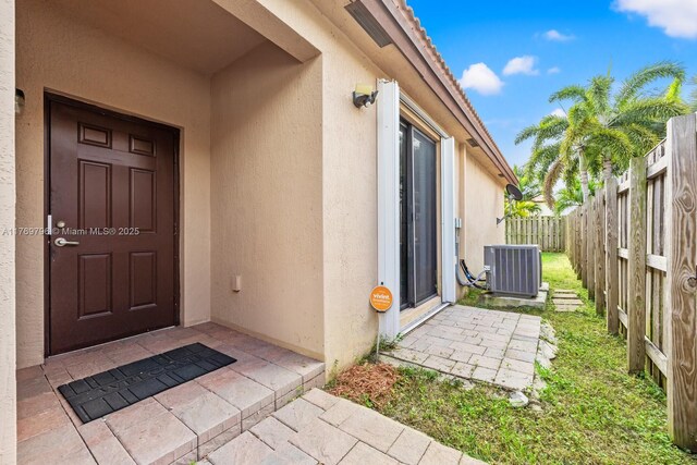 view of exterior entry with fence, stucco siding, and central air condition unit