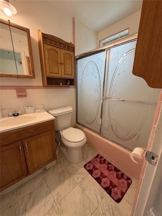 full bathroom with toilet, vanity, wainscoting, marble finish floor, and shower / bath combination with glass door