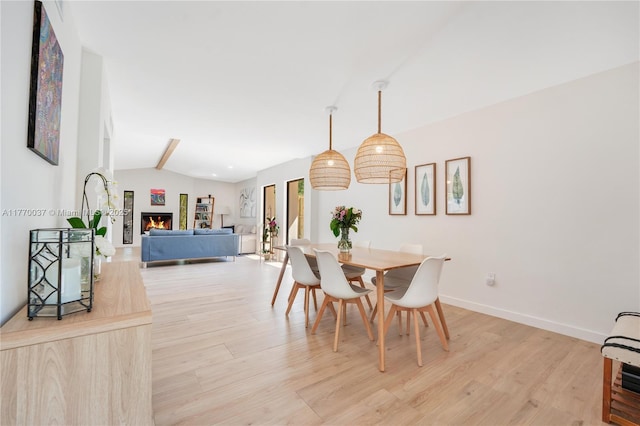 dining room featuring vaulted ceiling with beams, a lit fireplace, light wood-style flooring, and baseboards