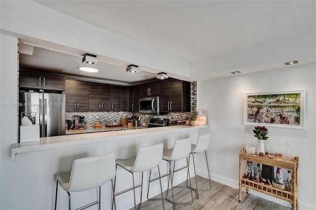 kitchen with light countertops, stainless steel appliances, dark brown cabinets, a breakfast bar area, and backsplash