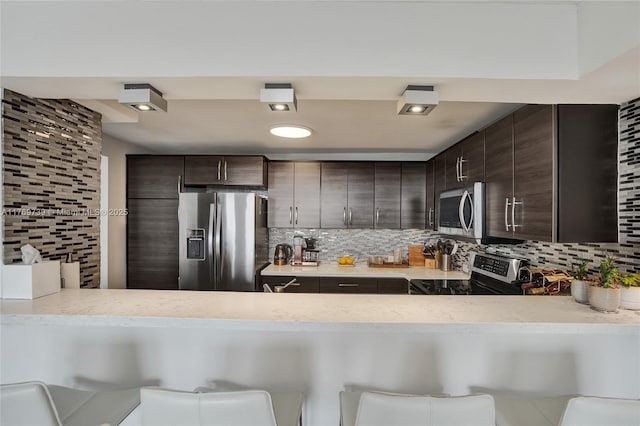 kitchen with dark brown cabinetry, decorative backsplash, a peninsula, and appliances with stainless steel finishes