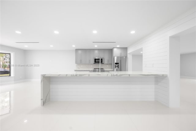 kitchen with tasteful backsplash, gray cabinetry, light tile patterned flooring, light stone countertops, and appliances with stainless steel finishes