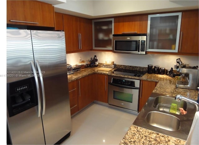 kitchen featuring dark stone counters, a sink, appliances with stainless steel finishes, brown cabinetry, and glass insert cabinets