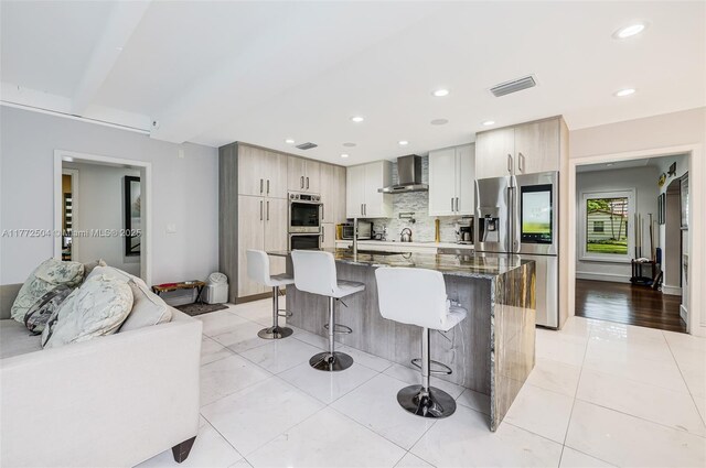 kitchen with a breakfast bar, dark stone counters, stainless steel appliances, wall chimney range hood, and backsplash