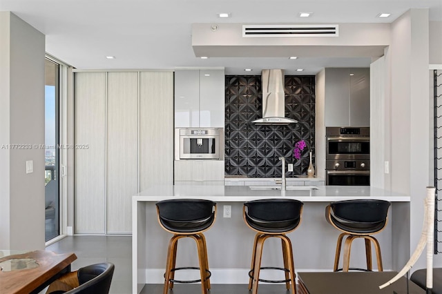 kitchen with wall chimney exhaust hood, a sink, modern cabinets, a breakfast bar, and double oven