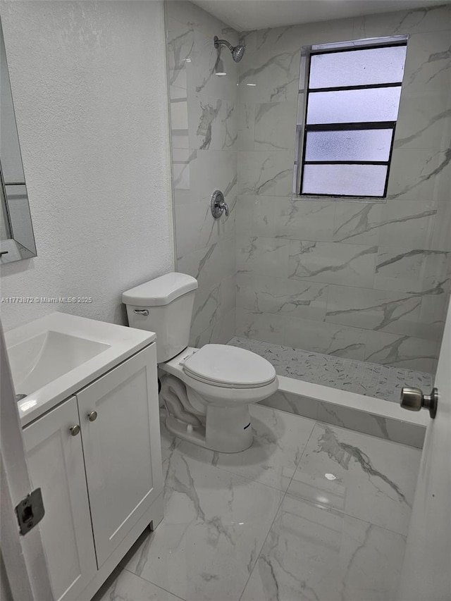 full bath featuring tiled shower, vanity, toilet, marble finish floor, and a textured wall