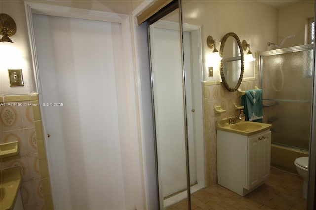 bathroom with toilet, tile walls, enclosed tub / shower combo, wainscoting, and vanity