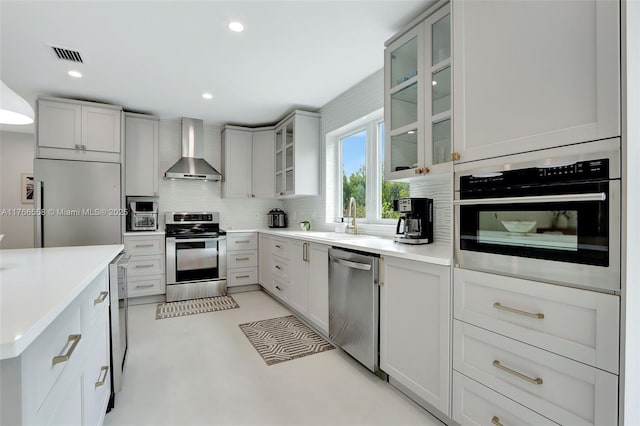 kitchen featuring glass insert cabinets, stainless steel appliances, wall chimney range hood, backsplash, and a sink