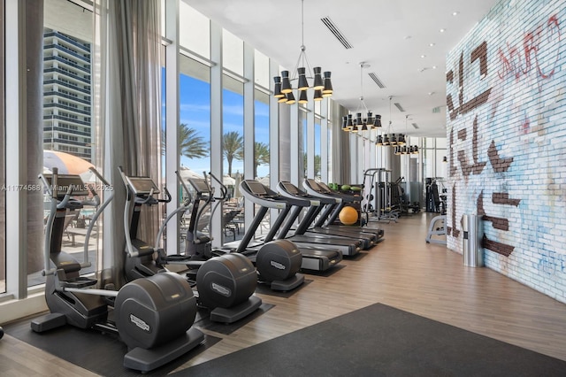 workout area with expansive windows, wood finished floors, brick wall, visible vents, and an inviting chandelier