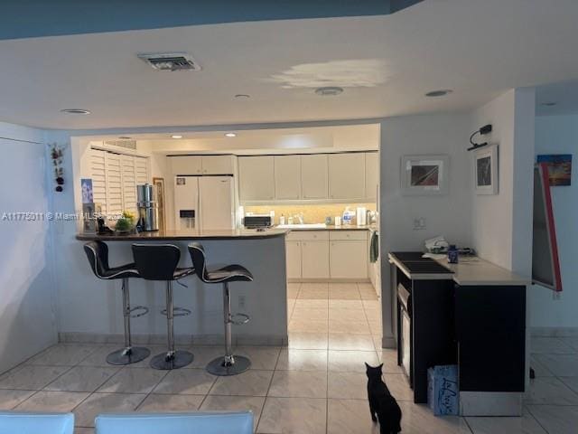 kitchen with a kitchen breakfast bar, a peninsula, white refrigerator with ice dispenser, white cabinets, and visible vents