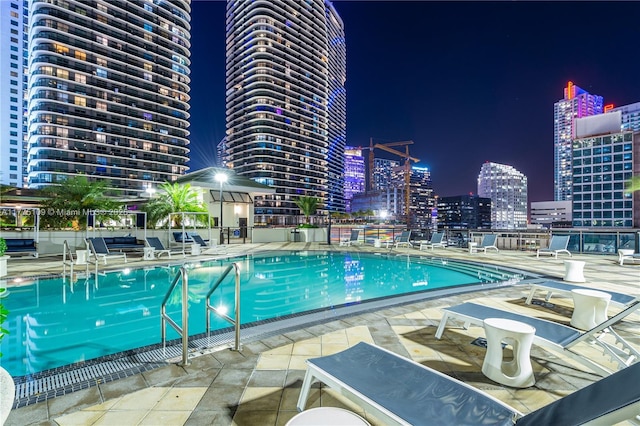 pool at twilight with a patio, fence, a view of city lights, and a community pool