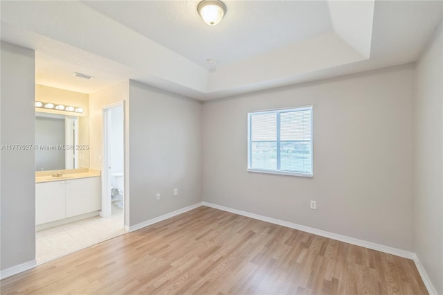 unfurnished bedroom featuring baseboards, a raised ceiling, a sink, light wood-type flooring, and connected bathroom