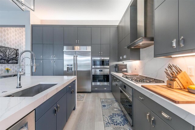 kitchen featuring tasteful backsplash, light wood-type flooring, stainless steel appliances, a sink, and wall chimney exhaust hood