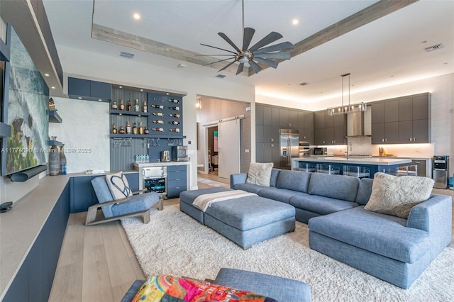 living area featuring a tray ceiling, a ceiling fan, a barn door, light wood-style flooring, and a dry bar