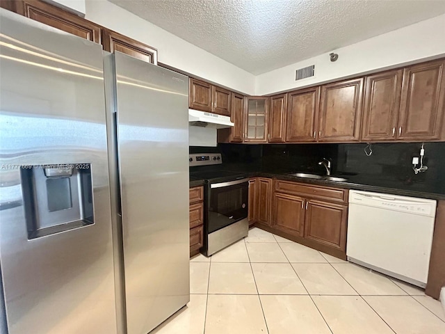 kitchen with dark countertops, appliances with stainless steel finishes, under cabinet range hood, a sink, and light tile patterned floors