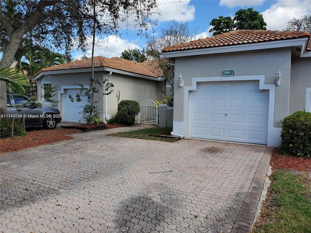 mediterranean / spanish-style home featuring a tiled roof, a garage, stucco siding, a gate, and decorative driveway