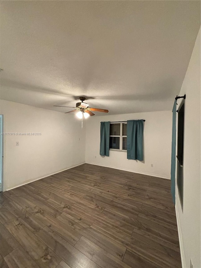 empty room with a barn door, a ceiling fan, a textured ceiling, dark wood finished floors, and baseboards