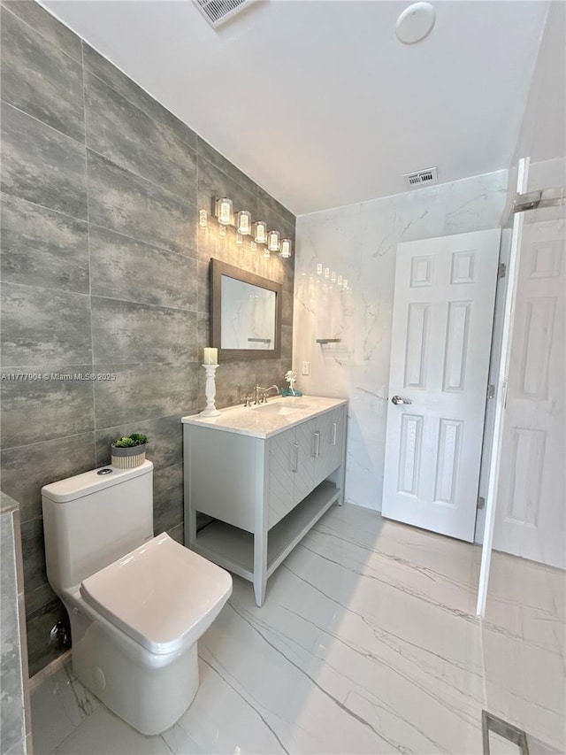 bathroom with toilet, vanity, marble finish floor, visible vents, and tile walls