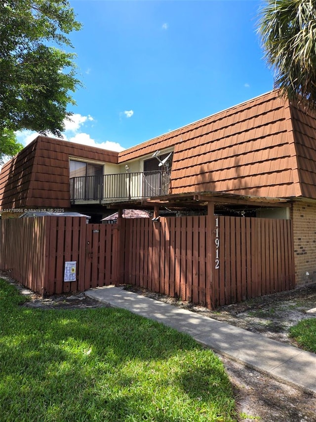 view of property exterior featuring fence, a balcony, mansard roof, and brick siding