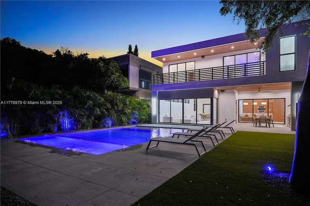 back of property at dusk with a lawn, a patio, stucco siding, an outdoor pool, and a balcony