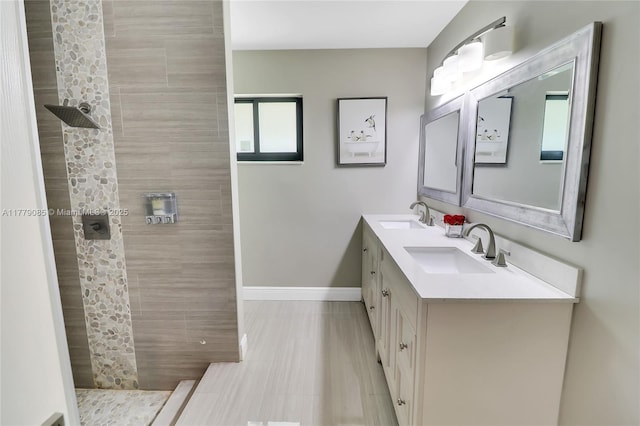 bathroom with tiled shower, double vanity, a sink, and baseboards