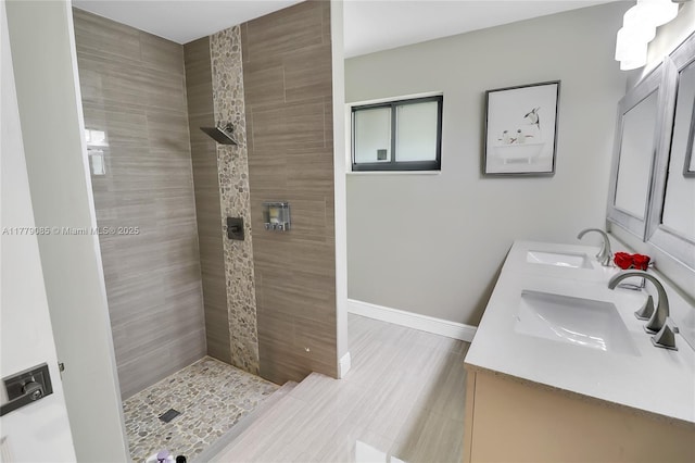 bathroom with tiled shower, double vanity, a sink, and baseboards