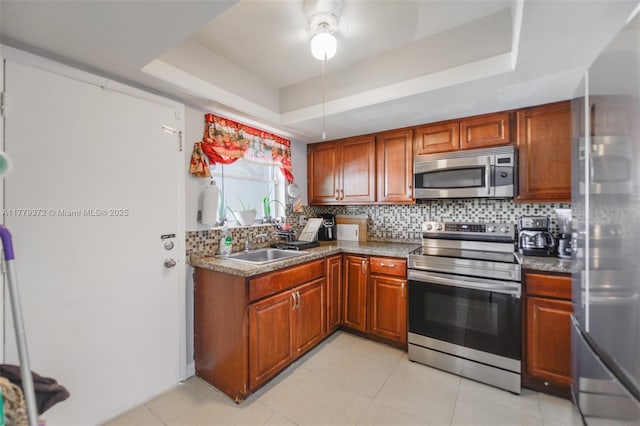 kitchen featuring brown cabinetry, a ceiling fan, backsplash, appliances with stainless steel finishes, and a tray ceiling