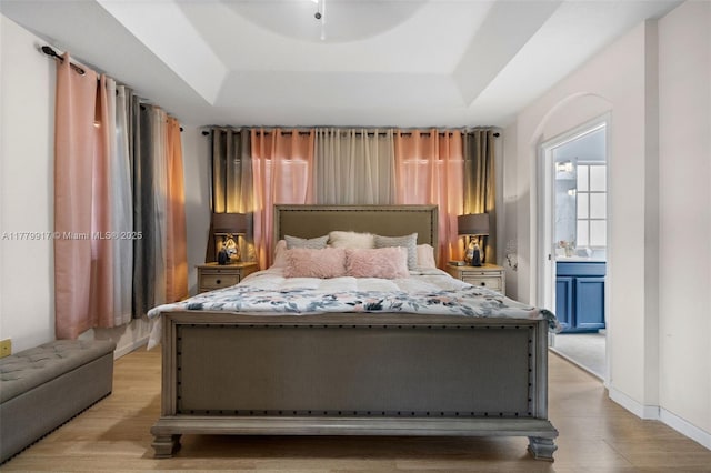 bedroom featuring ensuite bathroom, baseboards, a raised ceiling, and light wood finished floors