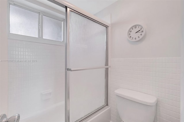 full bath featuring tile walls, a wainscoted wall, toilet, and combined bath / shower with glass door