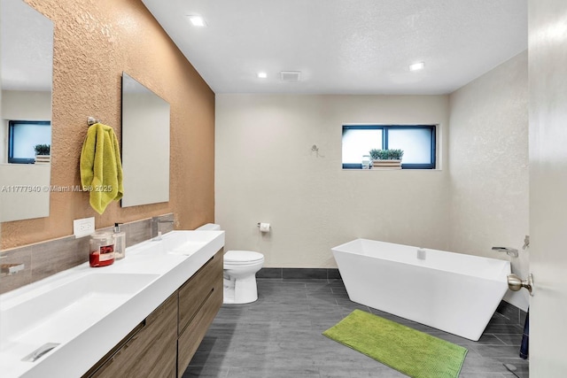 bathroom featuring a textured wall, a sink, double vanity, toilet, and a freestanding tub