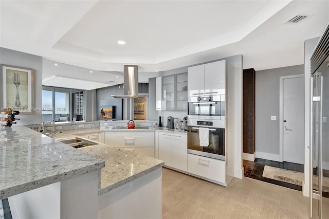 kitchen with modern cabinets, a tray ceiling, visible vents, light stone counters, and island range hood