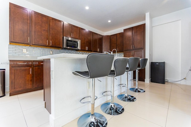 kitchen featuring light tile patterned floors, tasteful backsplash, stainless steel microwave, light countertops, and a breakfast bar area