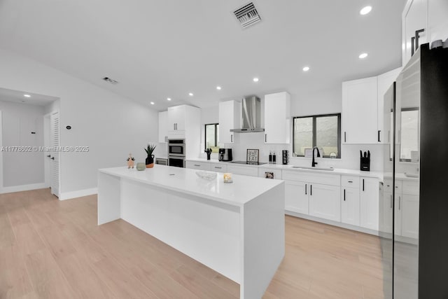 kitchen featuring wall chimney range hood, visible vents, a kitchen island, light countertops, and a sink