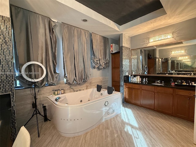 full bath with vanity, a tub with jets, a raised ceiling, tile walls, and wood finished floors
