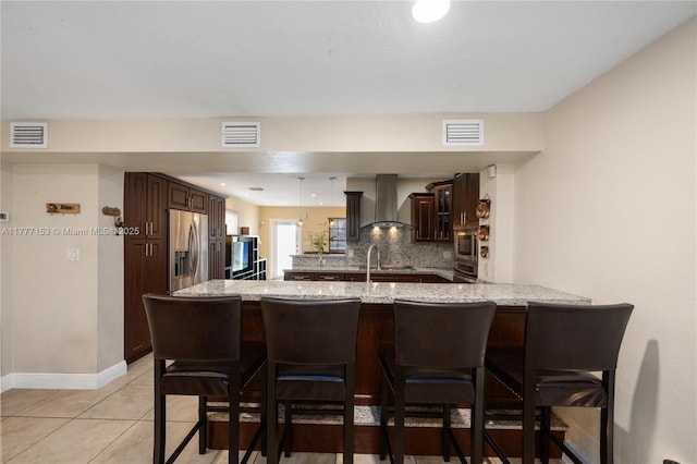 kitchen featuring a kitchen breakfast bar, wall chimney range hood, a peninsula, stainless steel refrigerator with ice dispenser, and visible vents