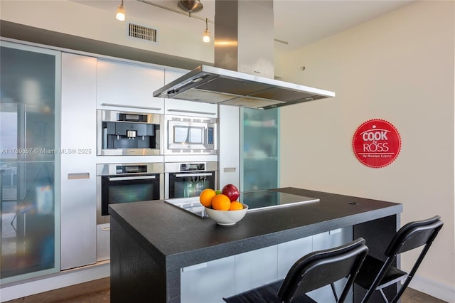 kitchen with track lighting, dark countertops, a breakfast bar area, island range hood, and stainless steel appliances