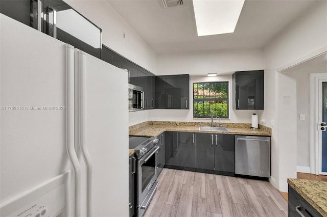 kitchen with light wood-type flooring, light stone counters, a sink, stainless steel appliances, and dark cabinetry