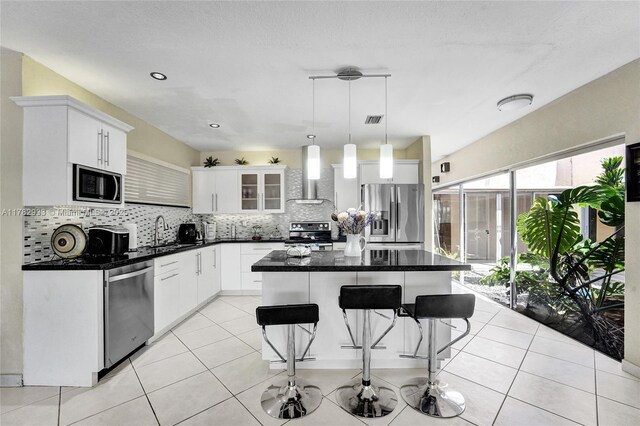 kitchen with wall chimney exhaust hood, dark countertops, appliances with stainless steel finishes, a kitchen bar, and backsplash