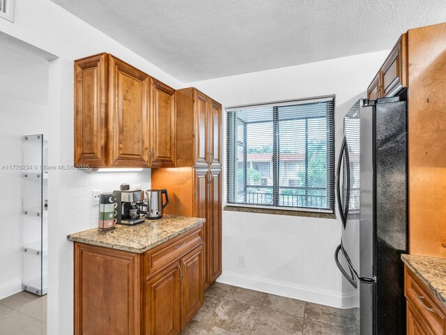 kitchen with brown cabinetry, freestanding refrigerator, baseboards, and light stone countertops