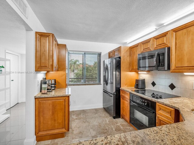 kitchen featuring tasteful backsplash, a textured ceiling, brown cabinets, black appliances, and light stone counters