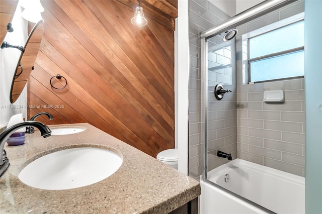 full bathroom with wooden walls, toilet, a sink, combined bath / shower with glass door, and double vanity