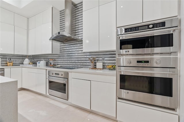 kitchen featuring wall chimney range hood, double oven, white cabinets, and black stovetop