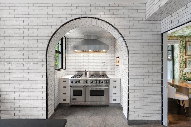 kitchen with light countertops, brick wall, wall chimney exhaust hood, and range with two ovens