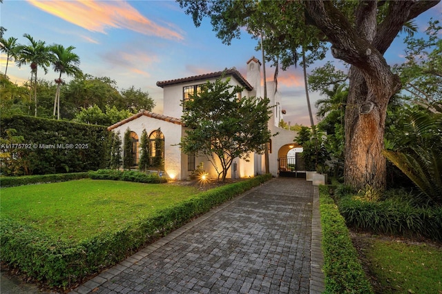 property exterior at dusk with stucco siding, a chimney, a tiled roof, a gate, and a lawn