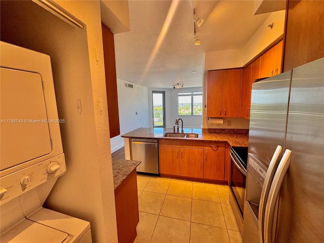 kitchen with stacked washer / drying machine, stainless steel appliances, a sink, a peninsula, and brown cabinetry