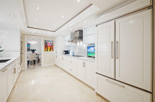 kitchen featuring a notable chandelier, a raised ceiling, wall chimney range hood, decorative backsplash, and black electric stovetop