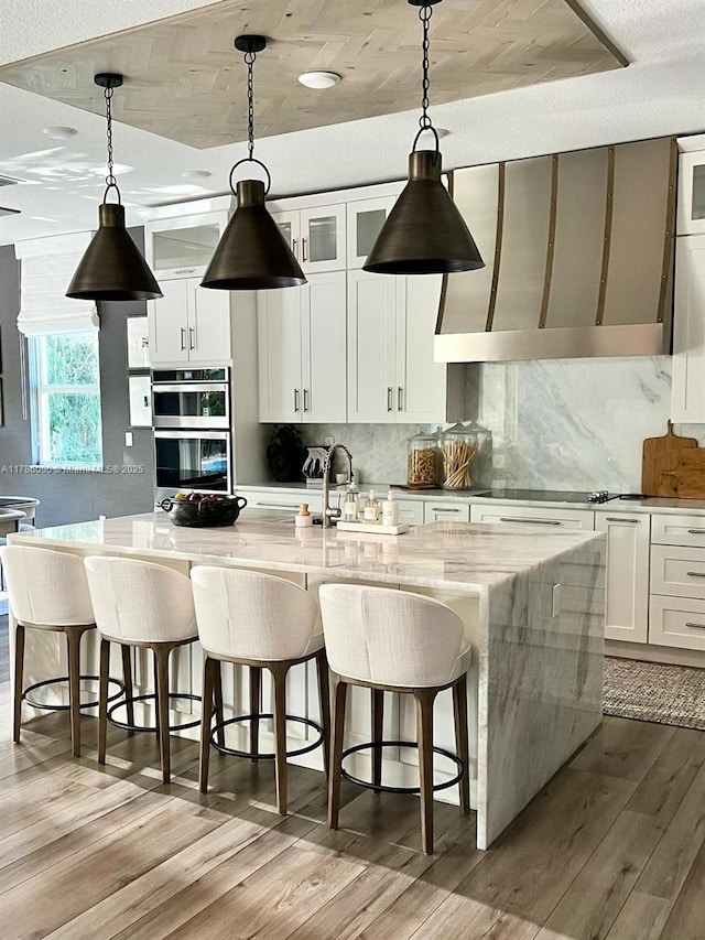 kitchen with white cabinets, double oven, backsplash, and a center island with sink